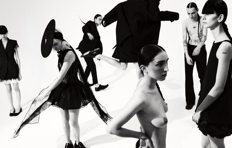 Fashion editorial shot of six slender models in a stark studio setting showcasing avant-garde black clothing, striking poses in a high-contrast black and white aesthetic.