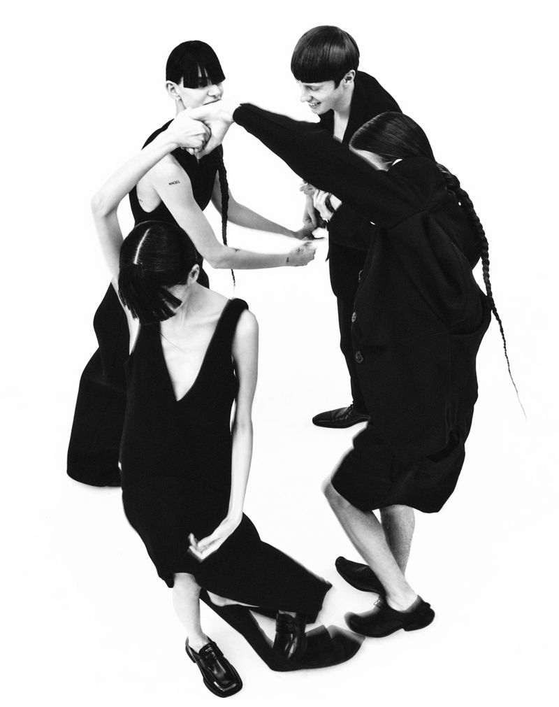 Black-and-white fashion shot featuring four young adult women in black dresses and matching shoes, in a bright studio setting, styled with modern haircuts.