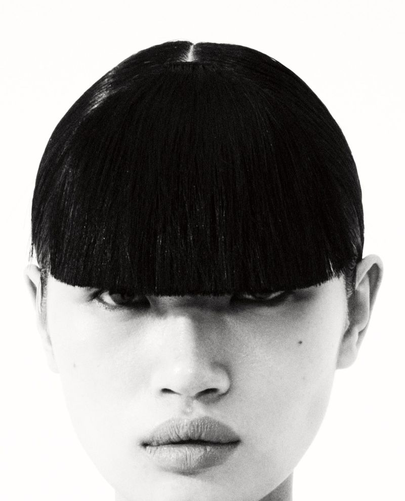 Close-up portrait of a woman with black bowl-cut hair, pale skin, and a neutral expression against a white background in a studio setting.