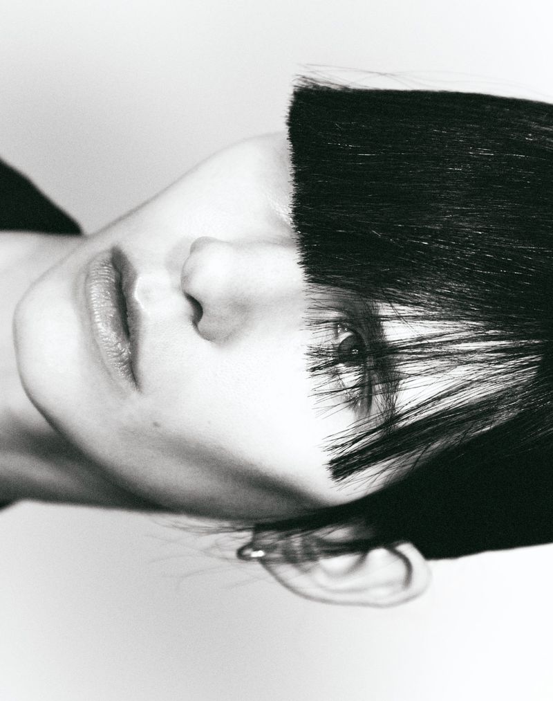 Monochrome close-up of an adult female's face with short black hair covering part of her face, facing forward with a neutral expression.