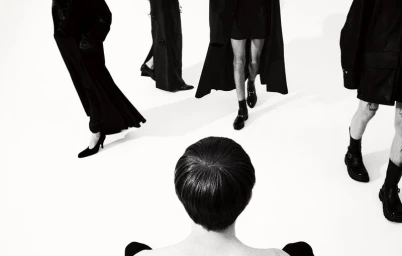 High-angle monochrome fashion editorial image featuring a seated person in the foreground observing models walking on a white studio floor.