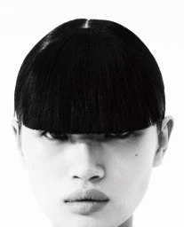 Close-up portrait of a model with short black blunt-cut bangs and direct gaze against a white background for Boycott Magazine.