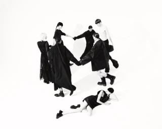Group of models in black clothing holding hands in a circle on a white background with one model lying on the floor