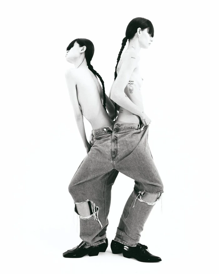Two models back to back wearing oversized distressed denim jeans and black shoes against a plain white background.