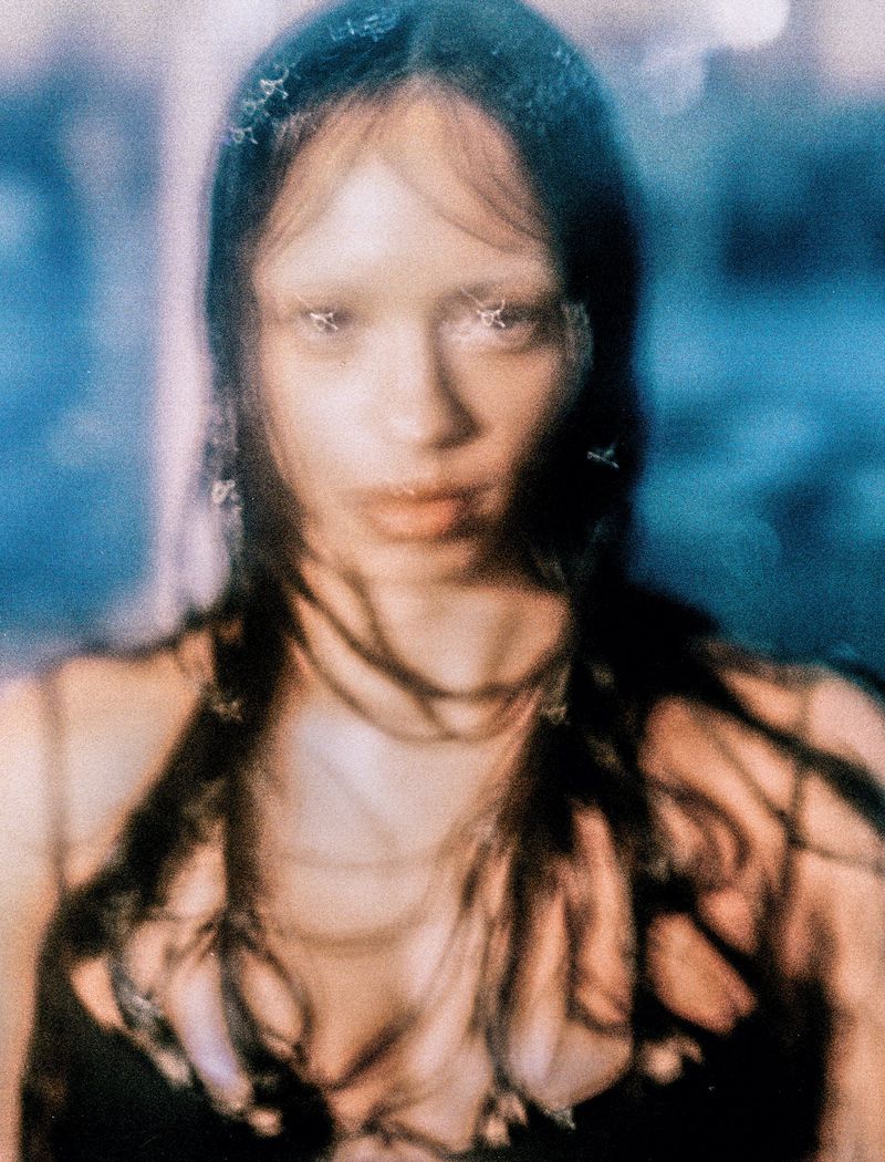 Close-up portrait of a young adult female with long dark hair, partially obscured by soft focus, set against a blue and peach-toned background.
