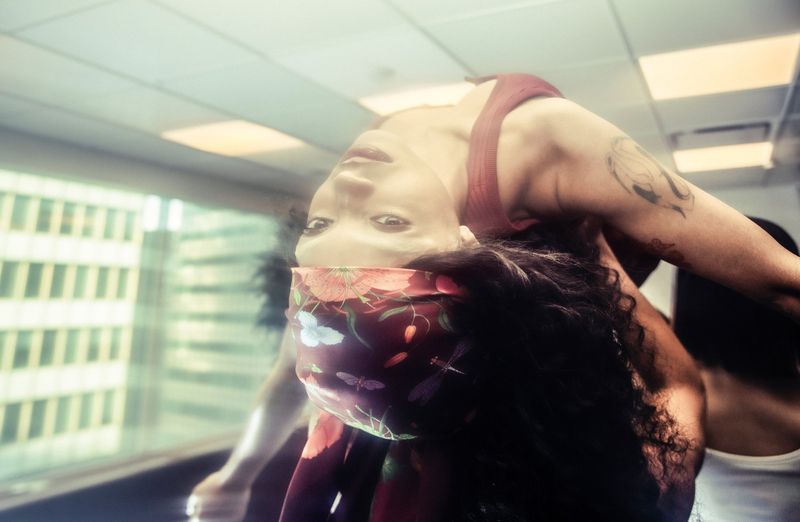 An adult woman arches back gracefully in a red top and patterned headband, with cityscape views through the window in the background.