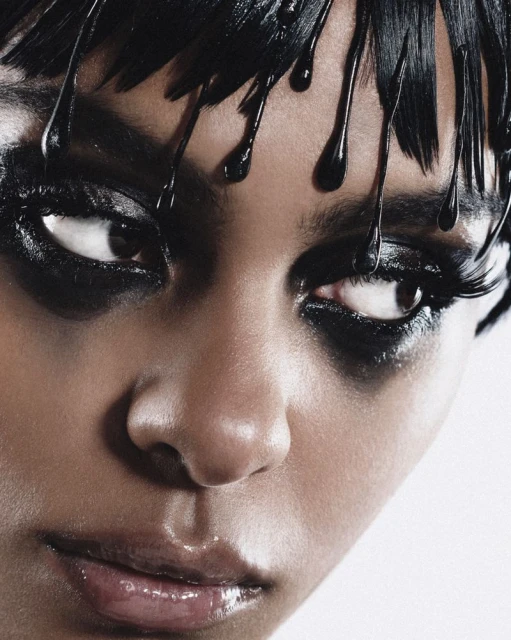 Close-up portrait of a woman with dark hair and striking black makeup around her eyes, set against a bright white backdrop.