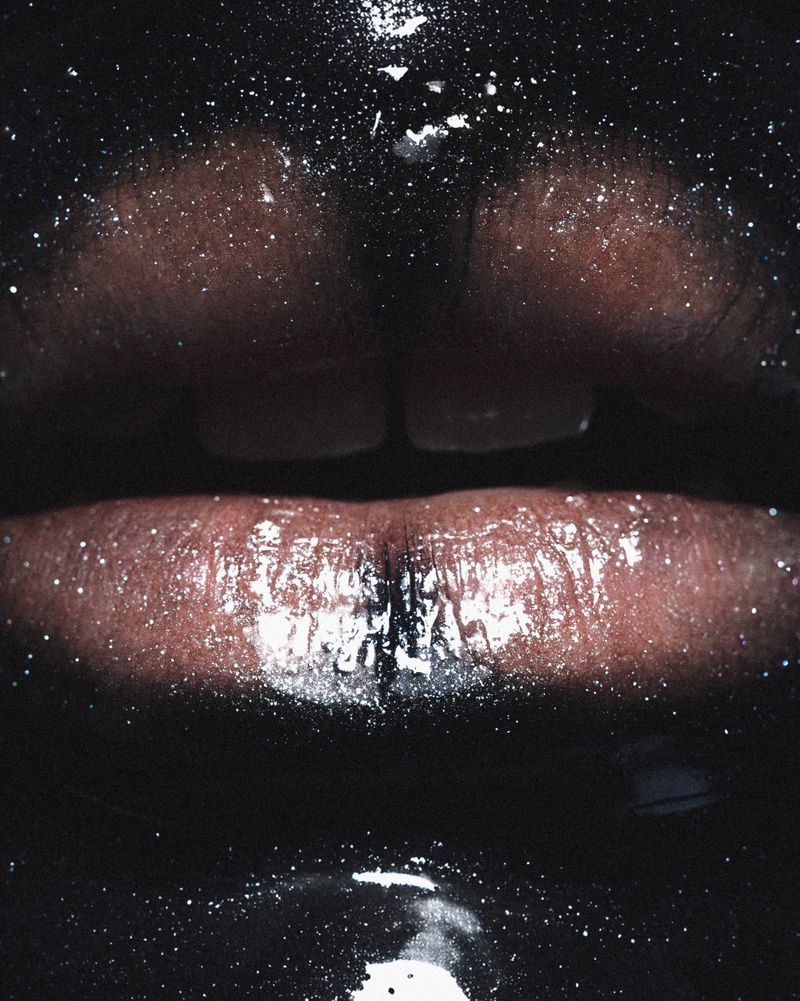 Close-up of dark-skinned person's glossy lips with glitter-like specks, set against a black background, highlighting a beauty or makeup aesthetic.