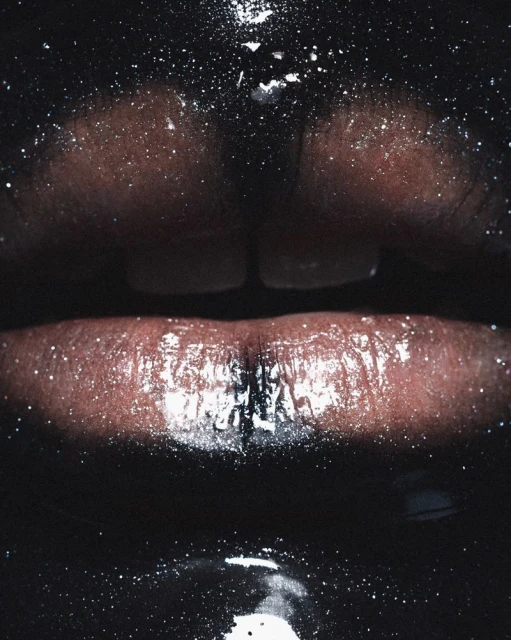 Close-up of dark-skinned person's glossy lips with glitter-like specks, set against a black background, highlighting a beauty or makeup aesthetic.
