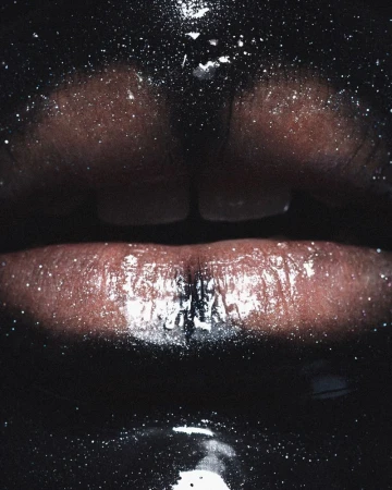 Close-up of dark-skinned person's glossy lips with glitter-like specks, set against a black background, highlighting a beauty or makeup aesthetic.