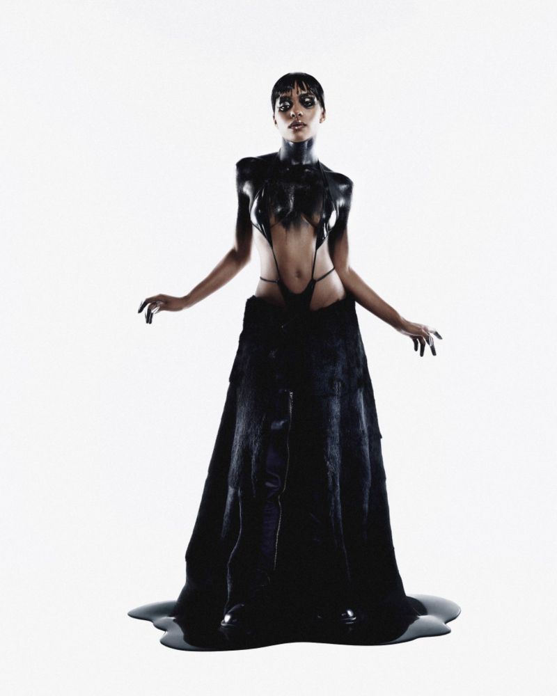 An adult woman poses in a unique black dress with dripping paint effect against a bright white studio backdrop, showcasing dark fashion.