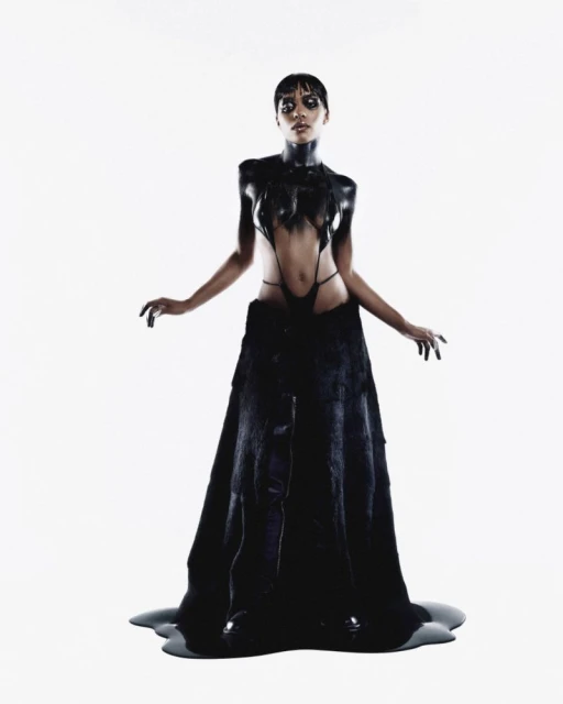 An adult woman poses in a unique black dress with dripping paint effect against a bright white studio backdrop, showcasing dark fashion.