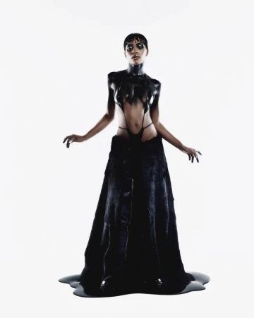 An adult woman poses in a unique black dress with dripping paint effect against a bright white studio backdrop, showcasing dark fashion.