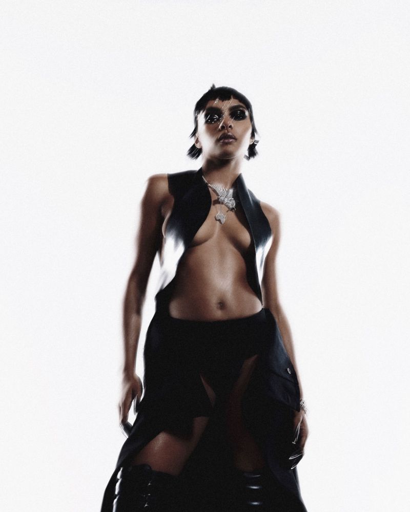 Woman posing in black vest and skirt with boots against a white background in a fashion-forward studio shot, wearing jewelry.