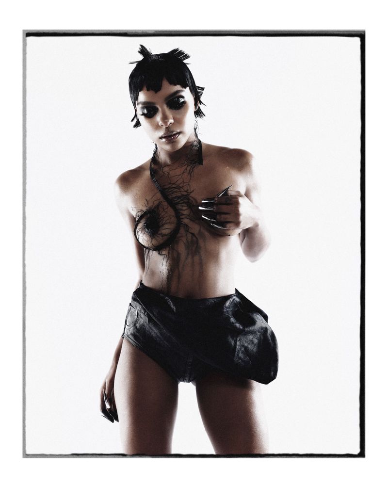 Artistic studio shot of a woman in minimal black attire, featuring unique hair and nail design, against a stark white backdrop.