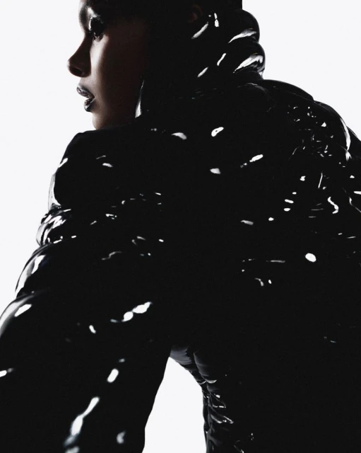 Profile portrait of a woman wearing a glossy black puffer jacket with a high collar, set against a bright white background in studio lighting.