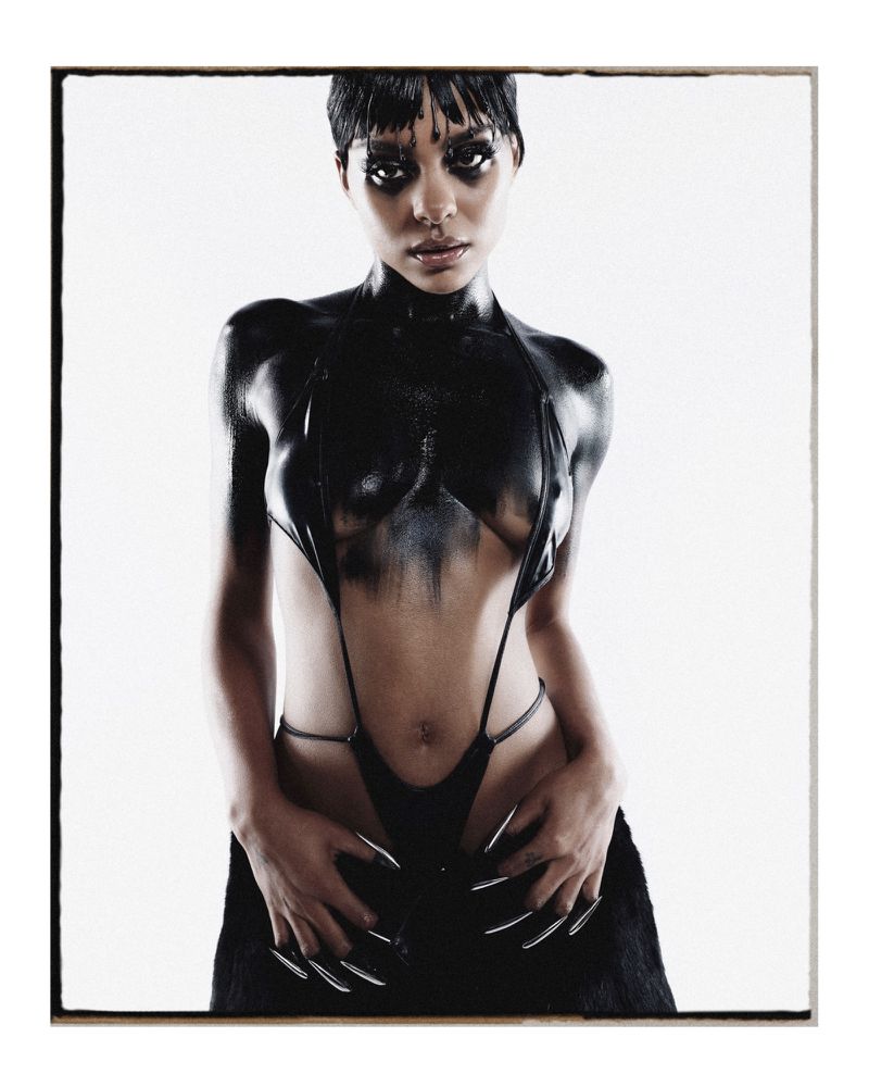 Striking portrait of a model in a glossy black body suit with long nails against a white background in a photo studio setting.