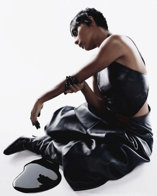 A woman in black leather clothing sits on a white surface next to a puddle of black liquid in a studio fashion shot.
