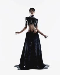 Model standing against a white seamless background wearing a black cutout top and long skirt with dark glossy body paint.
