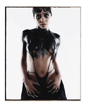 Portrait of a woman with dark hair and dramatic black body paint wearing a minimalist halter garment against a white background.