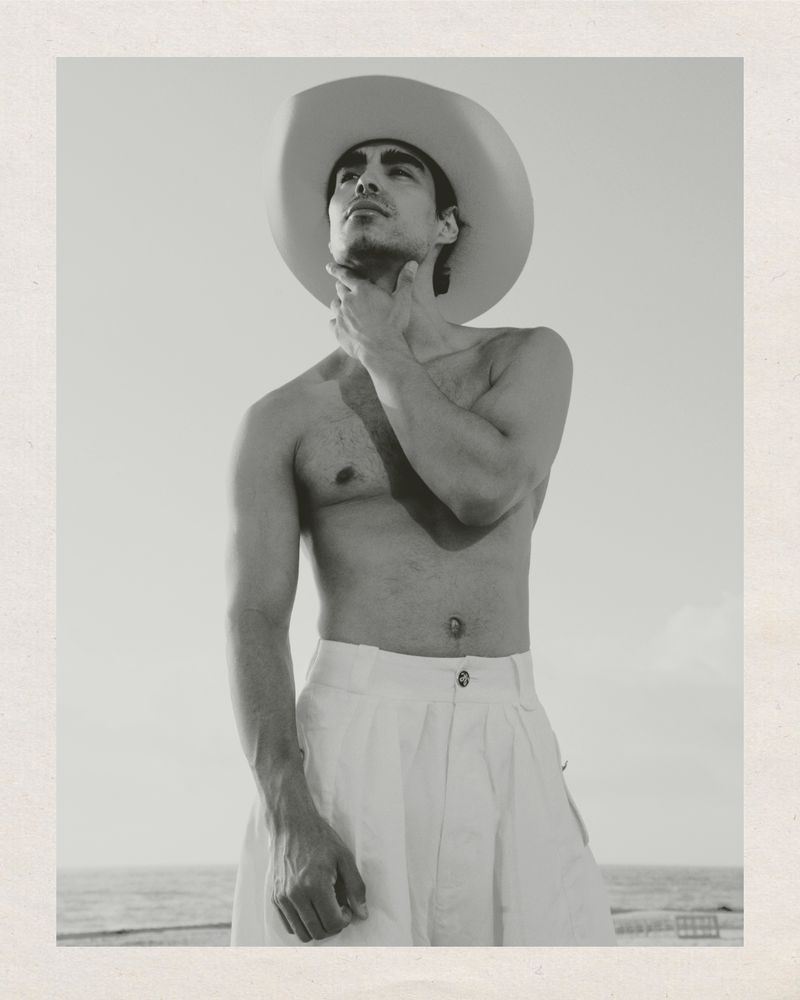 Monochrome portrait of a shirtless man in a white cowboy hat and pants, hand to chin, set against a sky and ocean backdrop.