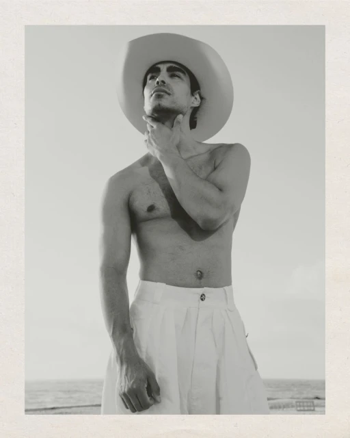 Monochrome portrait of a shirtless man in a white cowboy hat and pants, hand to chin, set against a sky and ocean backdrop.