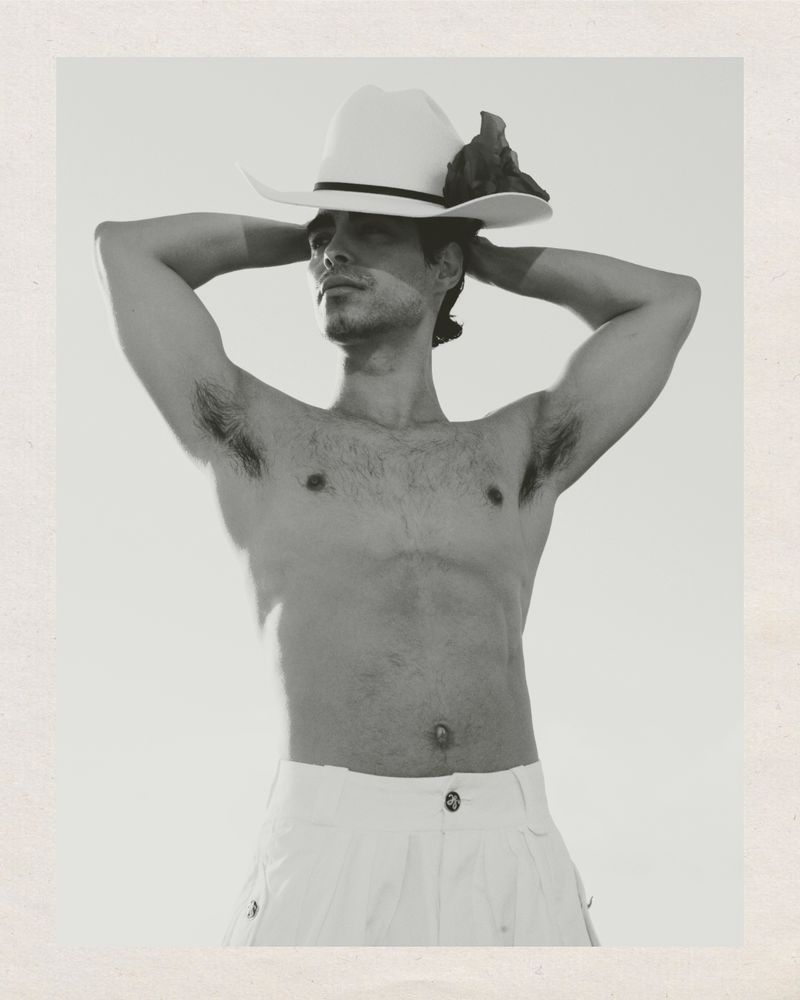 Monochrome portrait of a man wearing a cowboy hat and white pants, posing shirtless against a light background in a studio setting.