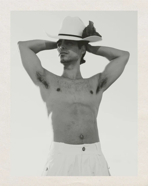 Monochrome portrait of a man wearing a cowboy hat and white pants, posing shirtless against a light background in a studio setting.