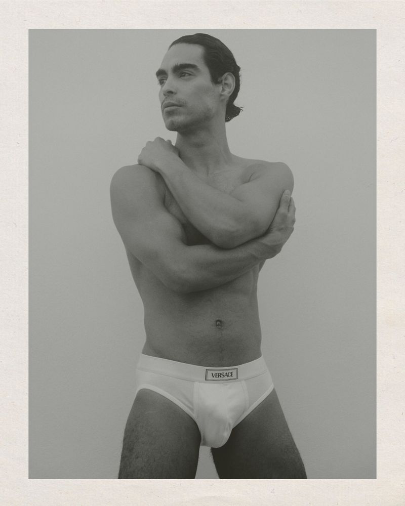 Monochrome studio portrait of a man posing in underwear, arms crossed, looking left with neutral facial expression against a plain backdrop.