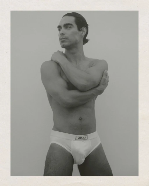 Monochrome studio portrait of a man posing in underwear, arms crossed, looking left with neutral facial expression against a plain backdrop.