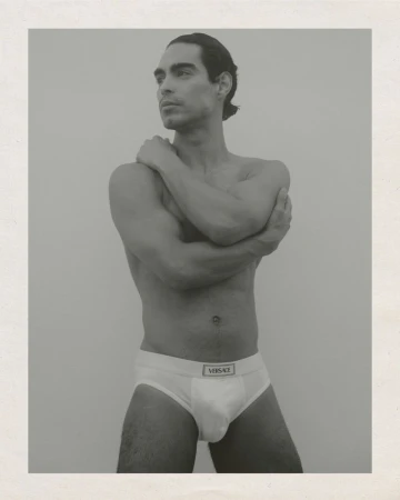 Monochrome studio portrait of a man posing in underwear, arms crossed, looking left with neutral facial expression against a plain backdrop.