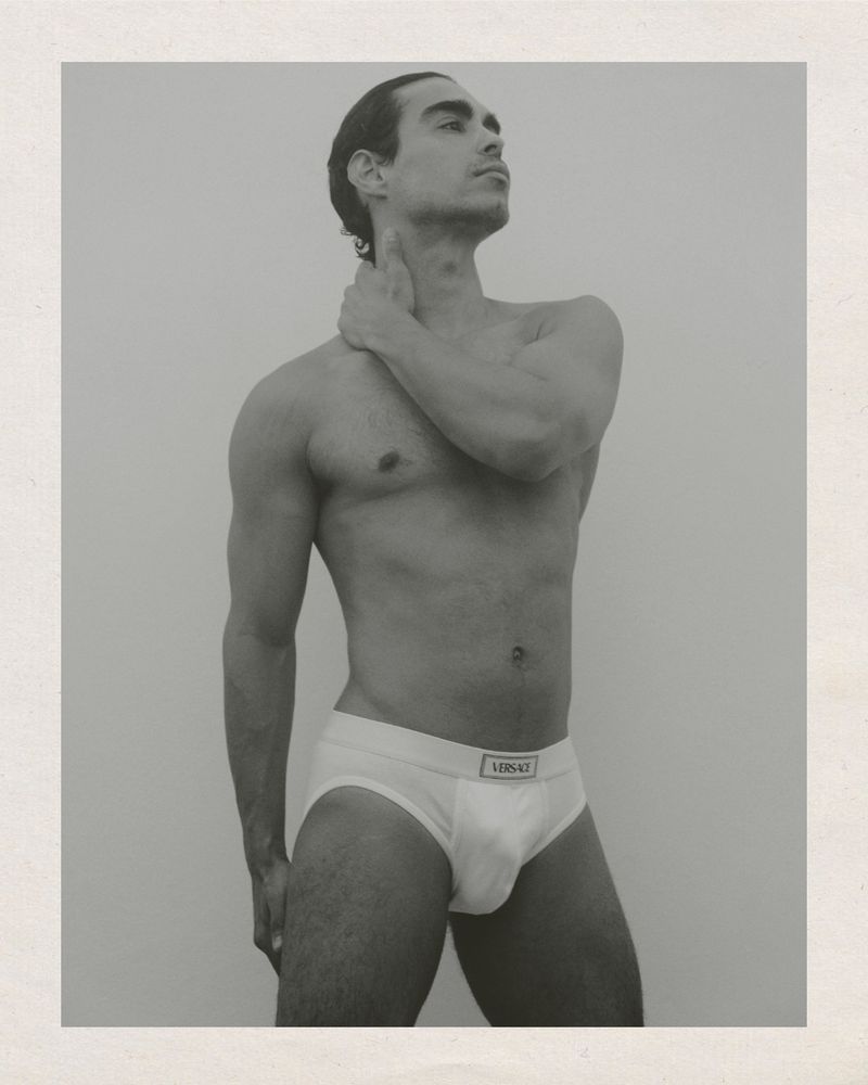 Monochrome portrait of a muscular adult male wearing white briefs with a logo, hand on neck, set against a neutral backdrop in studio light.