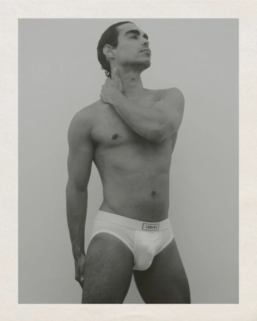 Monochrome portrait of a muscular adult male wearing white briefs with a logo, hand on neck, set against a neutral backdrop in studio light.
