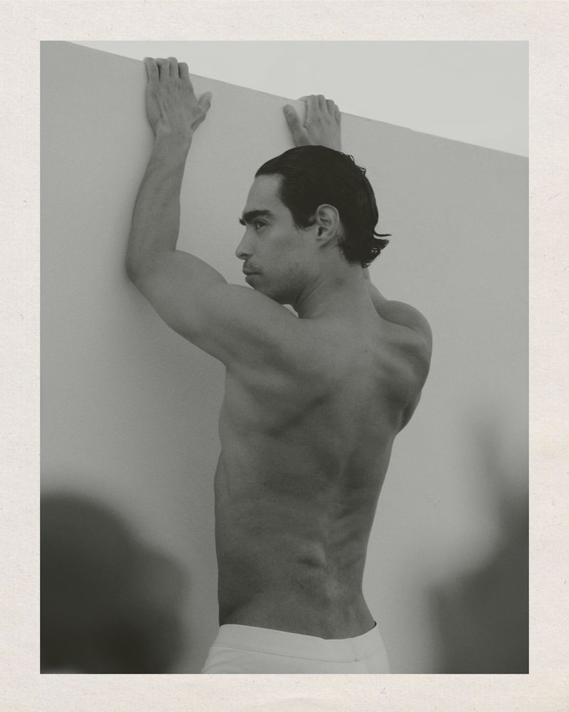 Monochrome portrait of a shirtless, athletic adult male leaning against a wall, showcasing his physique and dark hair in a studio setting.