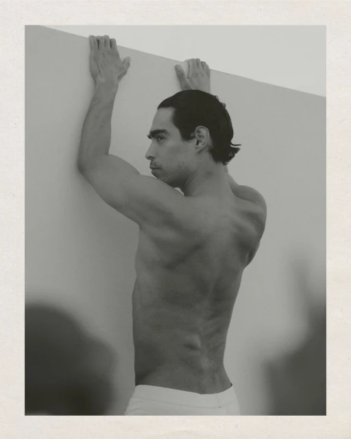 Monochrome portrait of a shirtless, athletic adult male leaning against a wall, showcasing his physique and dark hair in a studio setting.