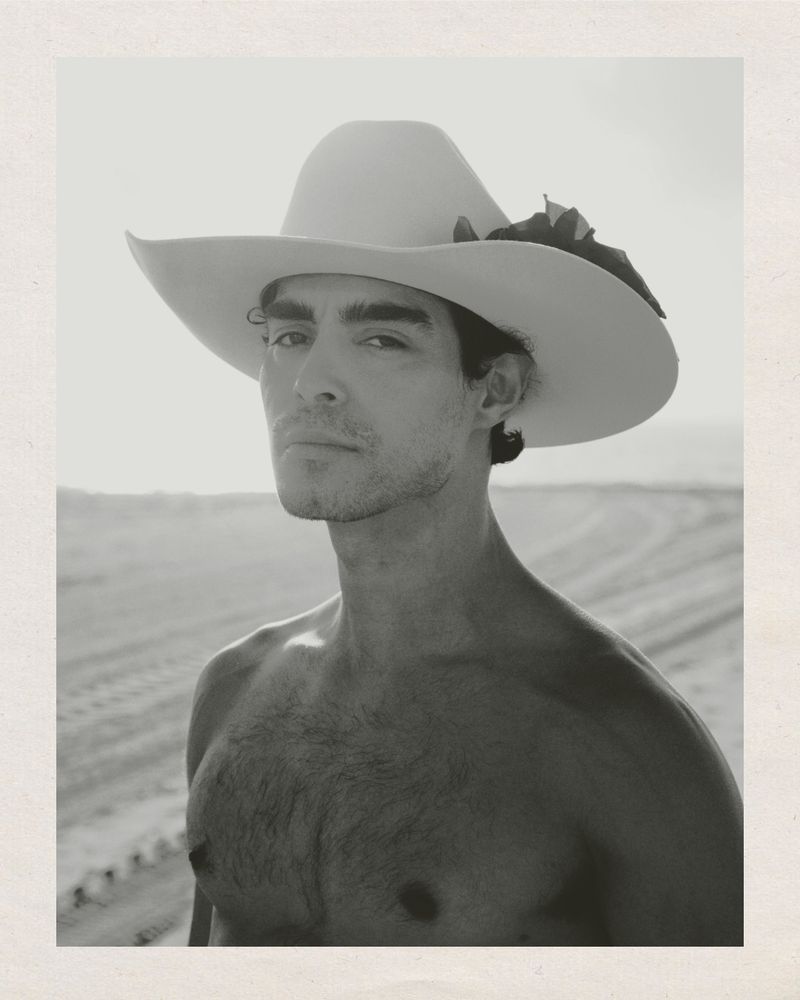 Black and white portrait of a man with a cowboy hat in a sandy environment. He has no shirt and is staring at the viewer.