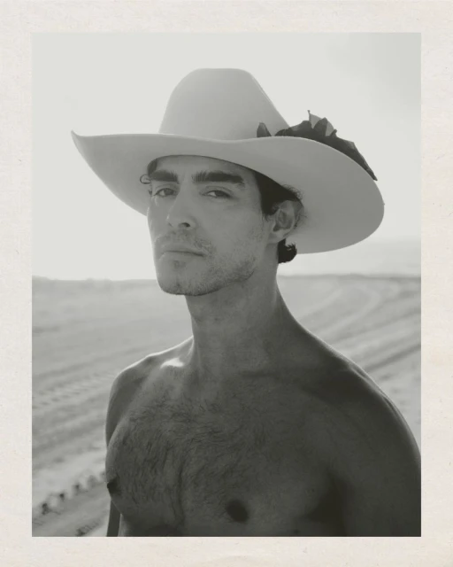 Black and white portrait of a man with a cowboy hat in a sandy environment. He has no shirt and is staring at the viewer.