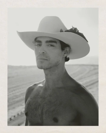 Black and white portrait of a man with a cowboy hat in a sandy environment. He has no shirt and is staring at the viewer.