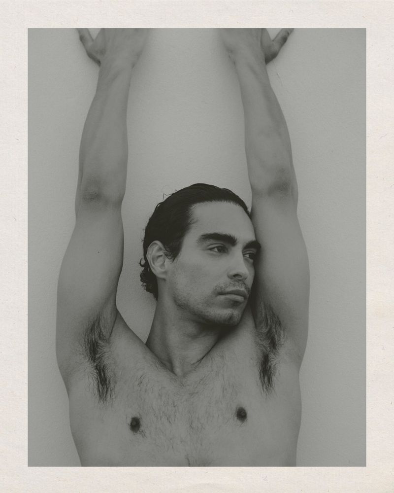 Monochrome studio portrait of a shirtless adult male with dark hair, looking off-camera, raising his arms against a light background.