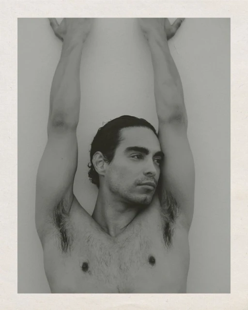 Monochrome studio portrait of a shirtless adult male with dark hair, looking off-camera, raising his arms against a light background.