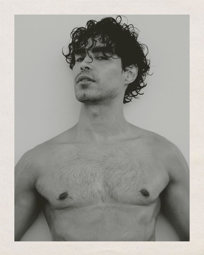 Black and white portrait of a shirtless man with curly dark hair looking upward against a plain background