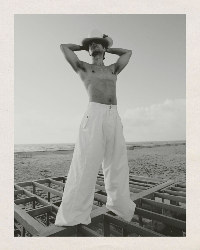 Shirtless man wearing wide-leg white trousers and a wide-brimmed hat standing on a wooden frame structure near the coast.