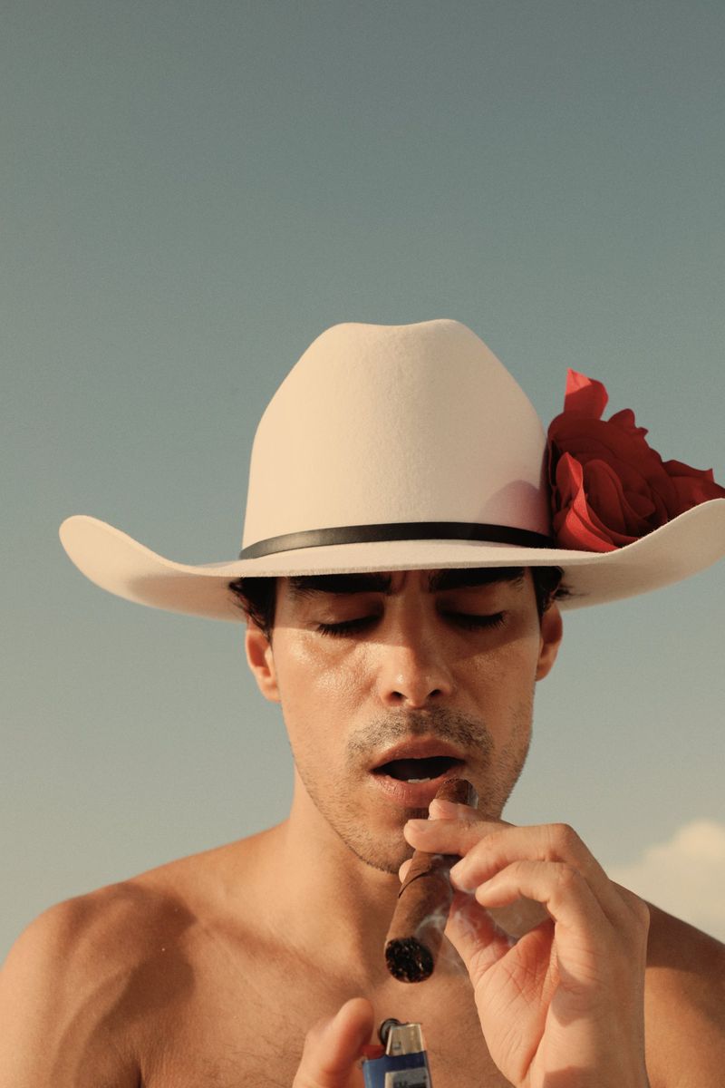 Adult male lighting cigar with a lighter, wearing a cowboy hat with a red rose, against a bright blue sky in an outdoor setting.