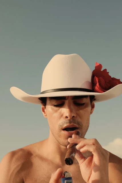 Adult male lighting cigar with a lighter, wearing a cowboy hat with a red rose, against a bright blue sky in an outdoor setting.