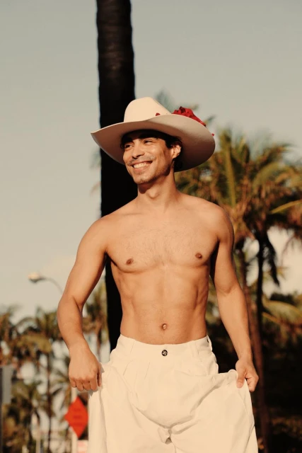 Shirtless man in white cowboy hat posing with palm trees in background. Male fitness, summertime tropical vibes, clear blue sky.