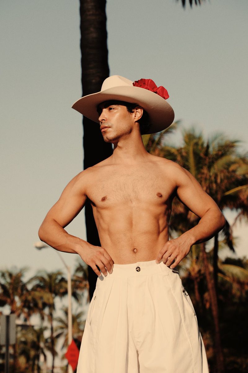 Shirtless man in wide-legged pants and cowboy hat poses outdoors with palm trees in background; fashion editorial, sunny daylight.