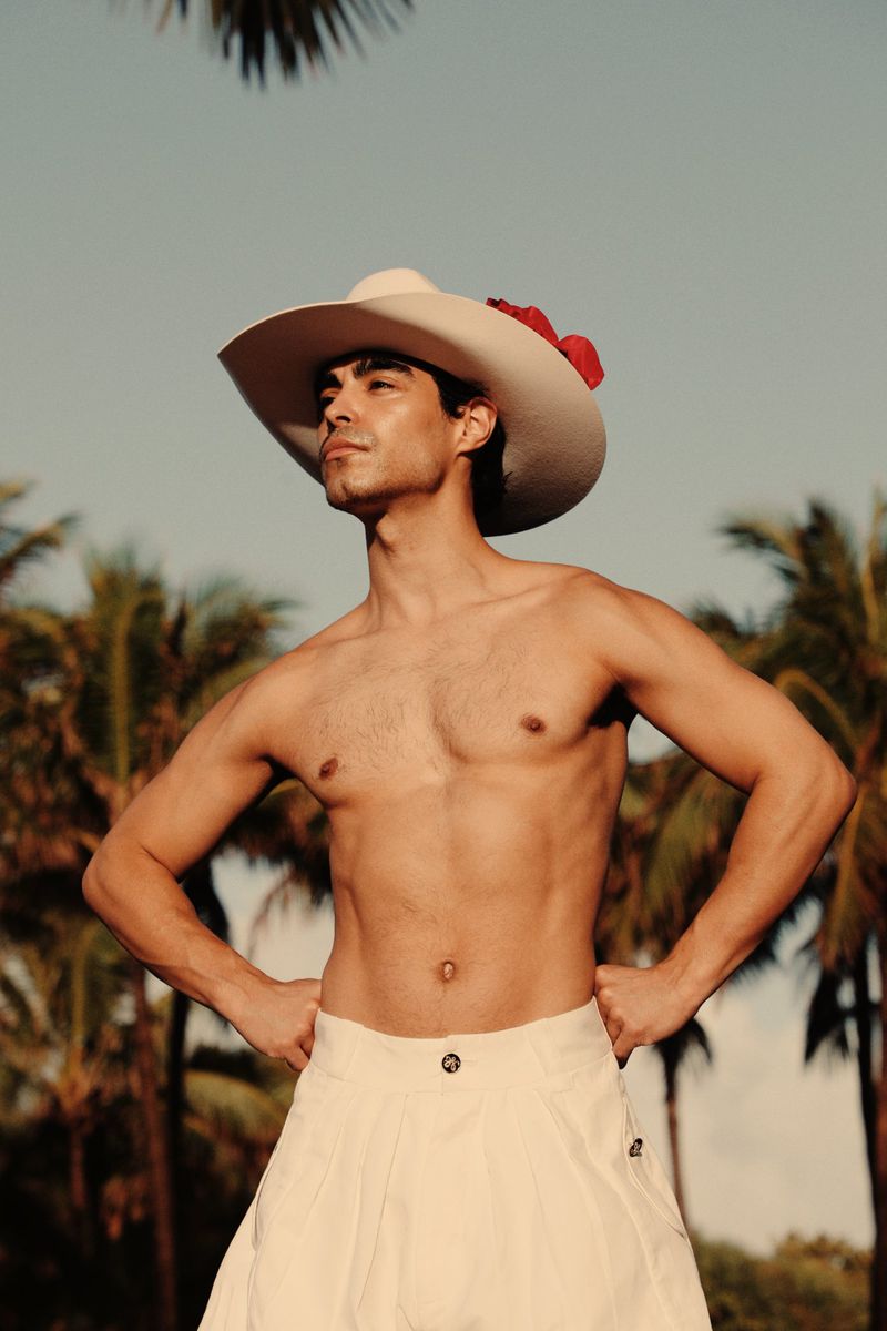 Shirtless man wearing white hat with red flower and white trousers standing against palm trees
