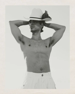Man wearing a white cowboy hat with hands behind head and white trousers against a light background