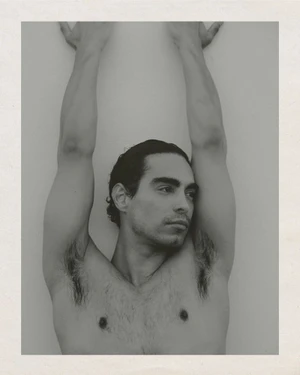 Portrait of a man with arms raised and hands resting against a plain white wall