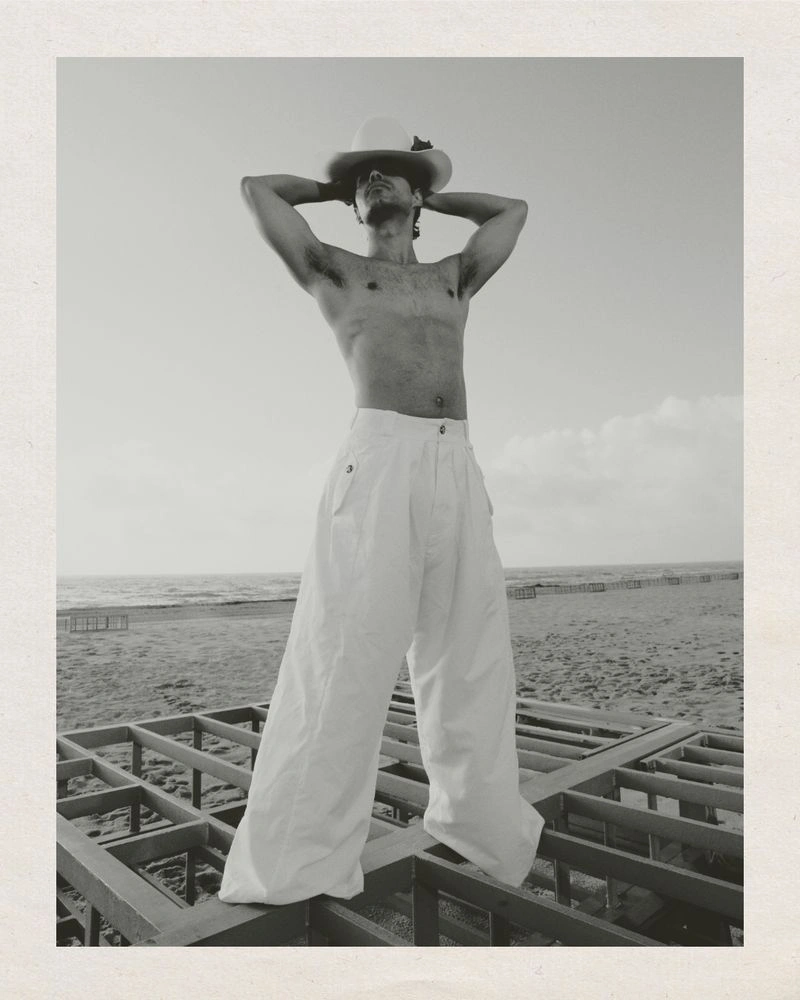 Shirtless man wearing wide-leg white trousers and a wide-brimmed hat standing on a wooden frame structure near the coast.